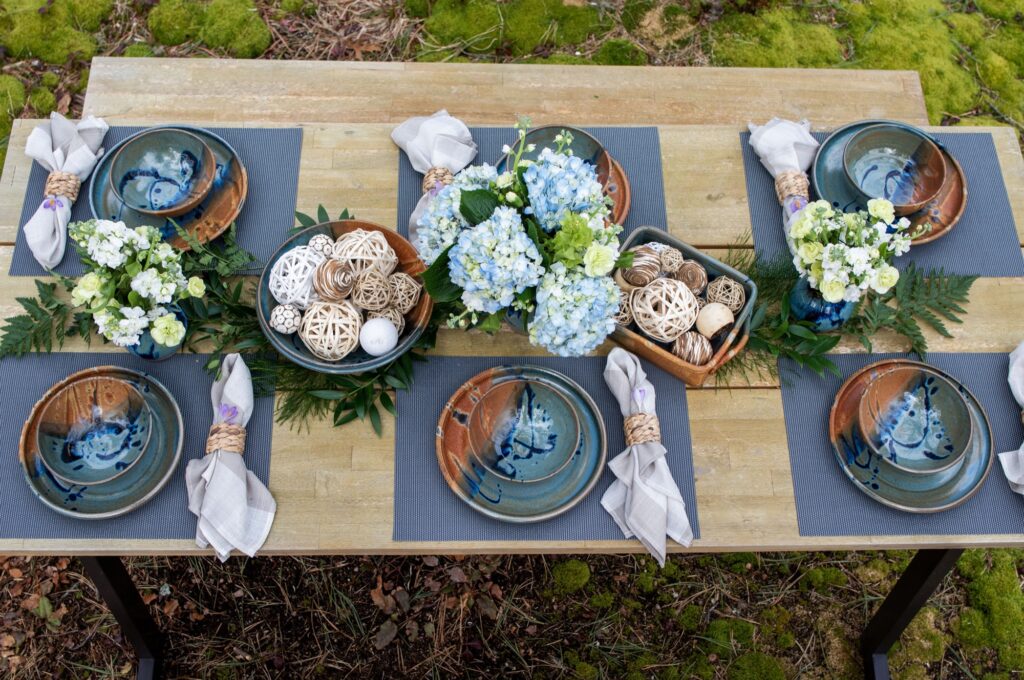 the photograph shows an overhead view of six place settings set up on a long rectangular wooden table. the table is set up outdoors, with a matching bench seen at the top edge of the photograph. each place setting consists of: a clay coyote soup and chili bowl, a clay coyote sandwich plate, a blue placemat, a linen napkin to the right of the plate and bowl, with a woven wicker napkin ring. each napkin and ring combo has a small purple flower tucked into it. down the middle of the table from left to right are as follows: fern branches, a small clay coyote vase with a white bouquet of flowers, more greenery, a clay coyote shallow salad bowl filled with white and brown decorative balls, a large bouquet of blue flowers, a clay coyote baking dish also filled with decorative white and brown balls, more greenery, a small clay coyote vase glazed in zappa with a white bouquet of flowers, and finally another fern branch. the photograph was taken outside from a overhead, top down viewpoint.