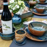 the photograph shows a close up of a tablescape made with clay coyote pottery. everything is on a light colored wooden table. starting from the left of the photograph working right there are: a clay coyote wine coaster, a yunomi, (both glazed in joes blue) a small vase glazed in zappa, a baking dish glazed in joes blue, a sandwich plate glazed in joes blue, with a joes blue soup and chili bowl resting on top of it and finally in the upper right corner, there is another sandwich plate and soup/chili bowl combo both done in joes blue. there are small branches of ferns decoratively laid on the table. each place setting has a blue placemat underneath the sandwich plate. the small zappa vase has a small bouquet of white flowers. the photograph was taken outside.