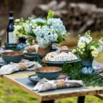 the photograph shows a picture from the side of a table set up outside filled with clay coyote pieces. the table has been set up for a dinner service, with multiple dinner place settings of dinner plates and soup bowls all glazed in joes blue. there is a large vase with a blue bouquet of flowers resting in it, the vase is glazed in joes blue. there is a deep salad bowl glazed in joes blue with multiple wicker balls decoratively placed within it. the wicker balls range from brown, to half brown half white to solid white. there is another bowl filled with these wicker balls, but it is mostly blocked from view by a soup and chili bowl, not allowing for identification. on the left side of the tablescape there is a bottle of wine, resting in a clay coyote wine coaster also in joes blue. there is a clay coyote yunomi near the wine bottle, also glazed in joes blue. each place setting has a linen napkin with a woven wooden napkin ring. each napkin ring has a small purple flower tucked into it. the photograph was taken outside with white natural light.