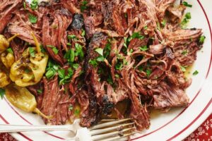 the photograph shows a white plate with red accents filled with shredded pot roast. there are pepperoncini peppers on the left side of the plate. the pot roast looks juicy and has liquid pooling ever so slightly on the bottom of the plate. there are fresh green herbs sprinkled over the shredded pot roast. at the bottom of the photograph and plate there are two silver colored forks resting with their tines pointed to the right. the forks are slightly overlapping, one on top of the other. in the bottom left and right corners, there is just a small peak of a red table cloth with small white flower designs.